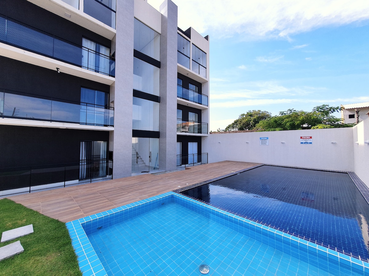 The pool area features two swimming pools, one with a vibrant blue tile design, surrounded by a wooden deck and greenery. The modern building facade stands in the background, showcasing large balconies and expansive glass doors that invite natural light.