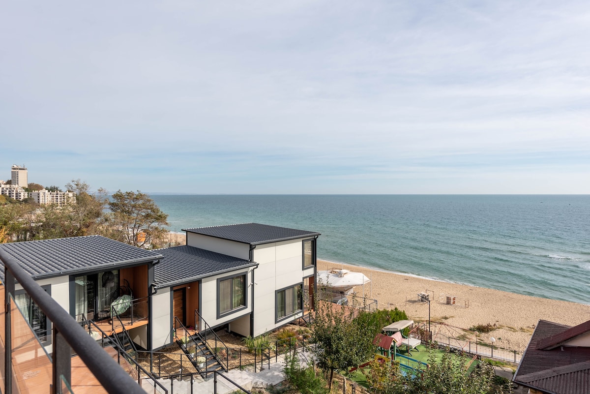 A panoramic view of the beach and ocean is captured from the balcony, highlighting the sandy shore and gentle waves. The two-story building is visible below, surrounded by greenery, with beach umbrellas in the distance.