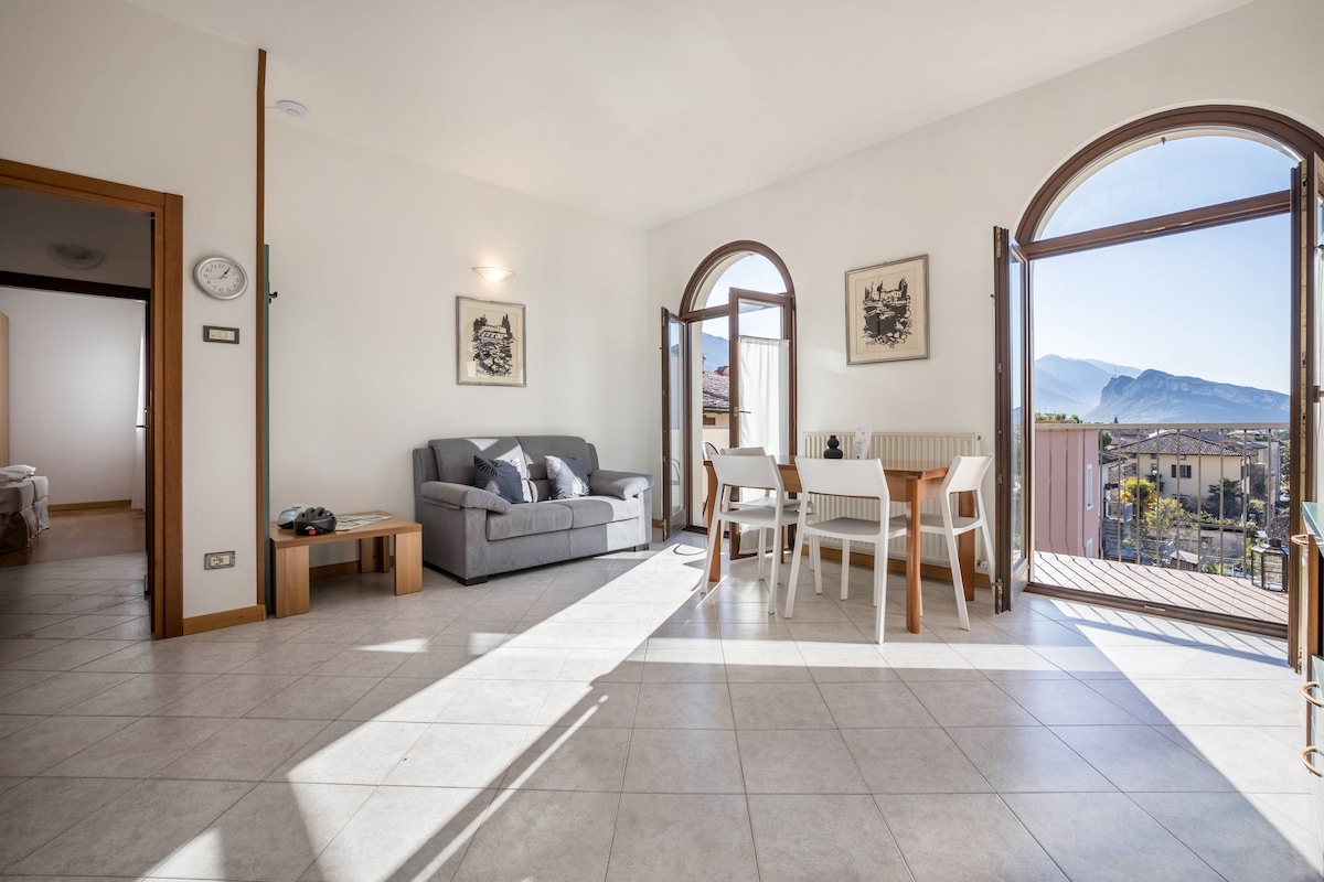 A bright living area features large arching windows, allowing natural light to flood the space. A cozy gray sofa and a wooden dining table with chairs are present, while tiled flooring extends beneath. Scenic mountain views are visible through the open doors, enhancing the ambiance.