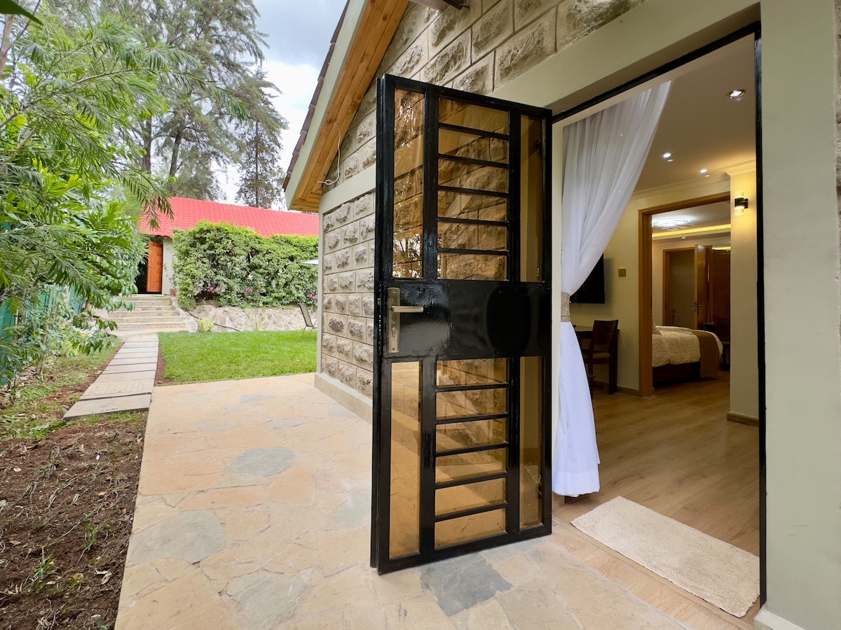 A black, gridded front door opens into a well-lit entryway. The stone path leads to a serene outdoor space surrounded by greenery, with a glimpse of a red-roofed structure in the background. Curtains sway gently in the entrance, inviting guests into the home.
