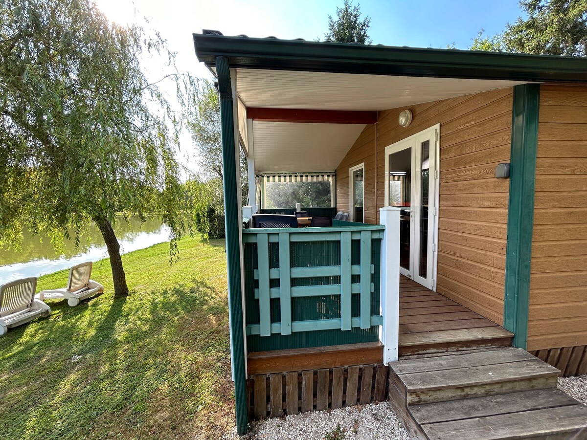 A cozy chalet exterior is visible, featuring a covered terrace with a railing. The pathway leads to a tranquil garden and an adjacent pond, enhancing the natural setting. Comfortable lounge chairs sit on the grass, inviting relaxation with beautiful views.