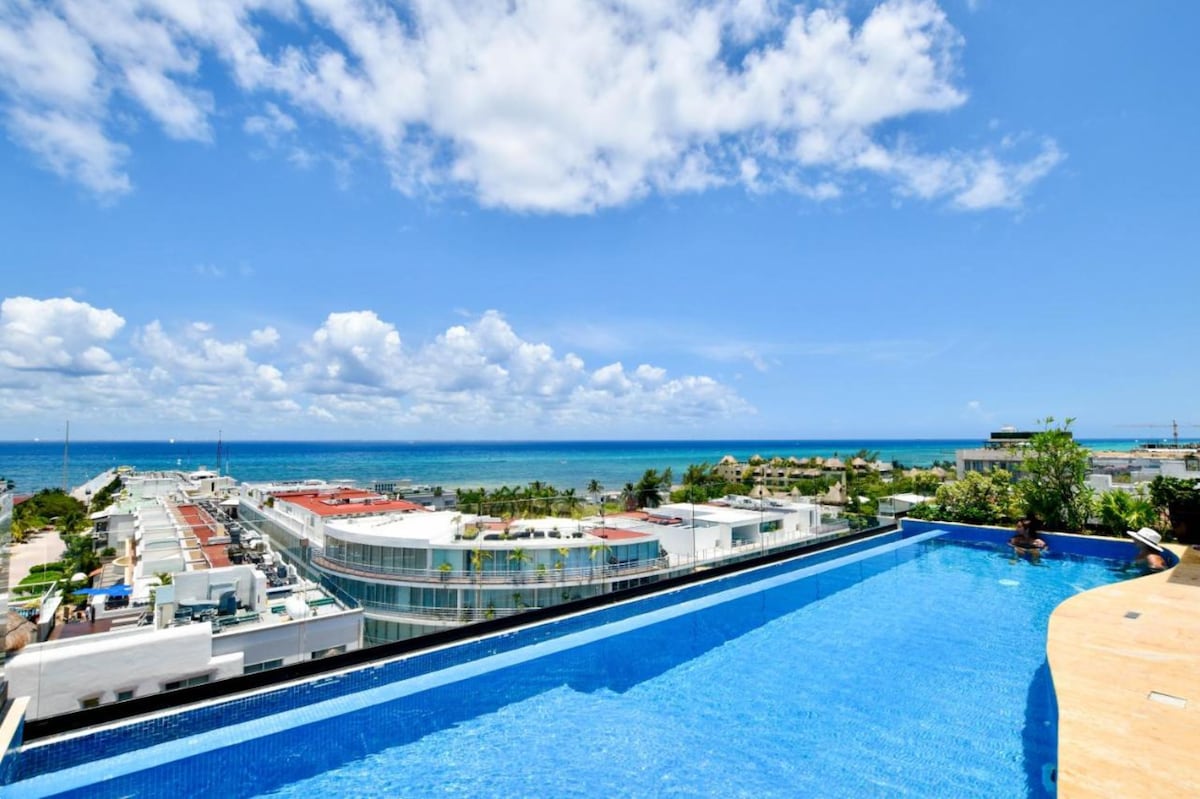 An expansive infinity pool extends towards the horizon, blending seamlessly with the turquoise sea. The vibrant sky is dotted with fluffy clouds, enhancing the picturesque setting. Nearby buildings and greenery contribute to the coastal view, creating a serene atmosphere.