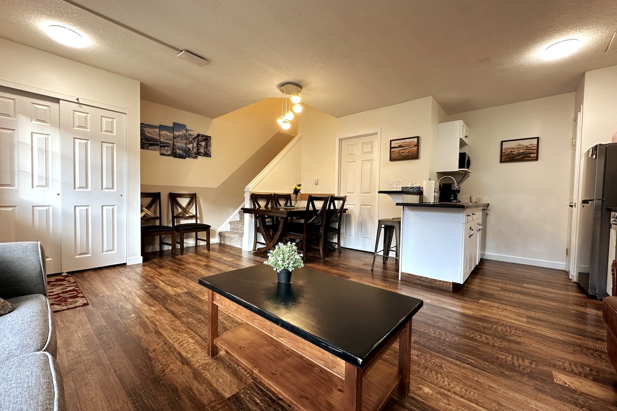 An inviting living area is furnished with a comfortable sofa and a coffee table. A dining table with six chairs is positioned nearby. The open kitchen features modern appliances, with light-colored cabinetry providing a bright atmosphere. Soft lighting and wooden flooring enhance the cozy ambiance.