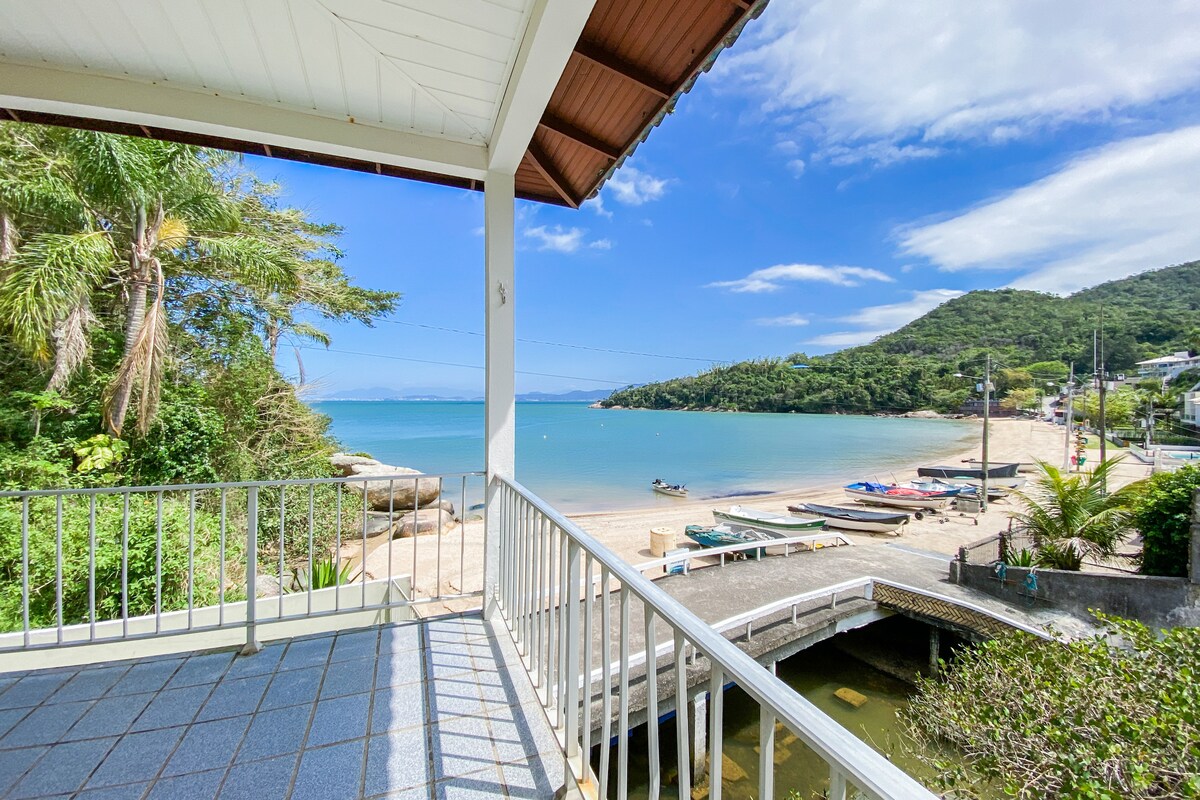 A balcony view showcases the shoreline with gentle waves lapping against the beach. Boats are moored nearby, and lush greenery lines the opposite shore, while a distant hill provides a scenic backdrop under a bright blue sky with scattered clouds.