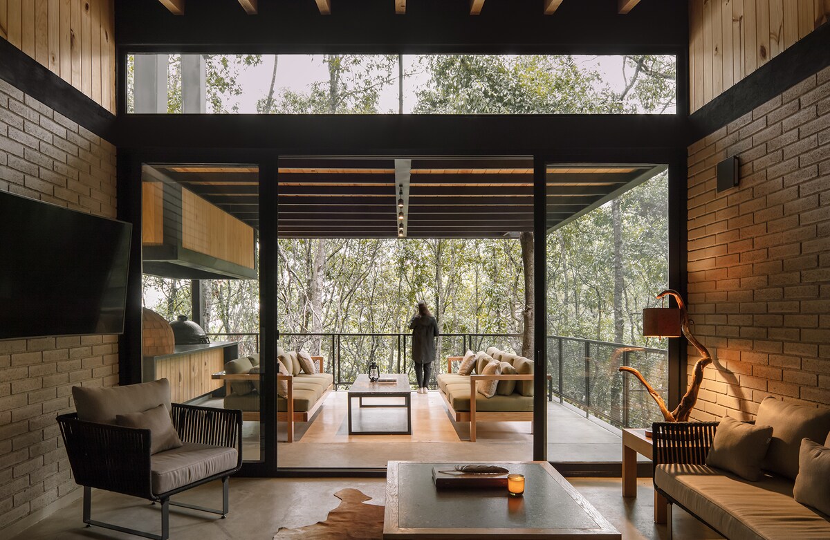 A spacious living area features large glass doors that open to a forest view. Natural wood and stone elements define the space, complemented by modern furnishings. A decorative lamp stands adjacent to the seating area, while a low coffee table sits centrally, enhancing the ambiance.