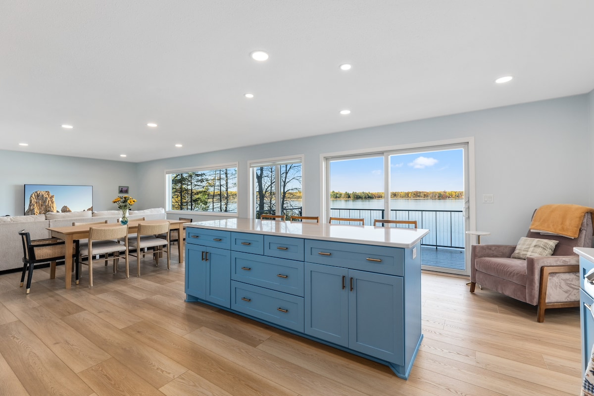 A bright and airy living space features a large blue kitchen island with ample storage. The open layout showcases a dining area and comfortable seating. Large windows provide natural light and views of the lake, creating a serene backdrop.