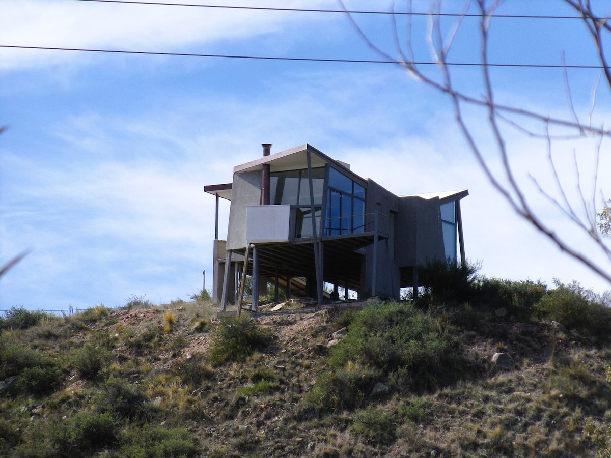 A modern house is situated on a hillside, elevated on stilts. Large windows and a sloped roof are prominent features, allowing for expansive views of the surrounding landscape. Natural vegetation is visible in the foreground, highlighting the home's connection to its environment.
