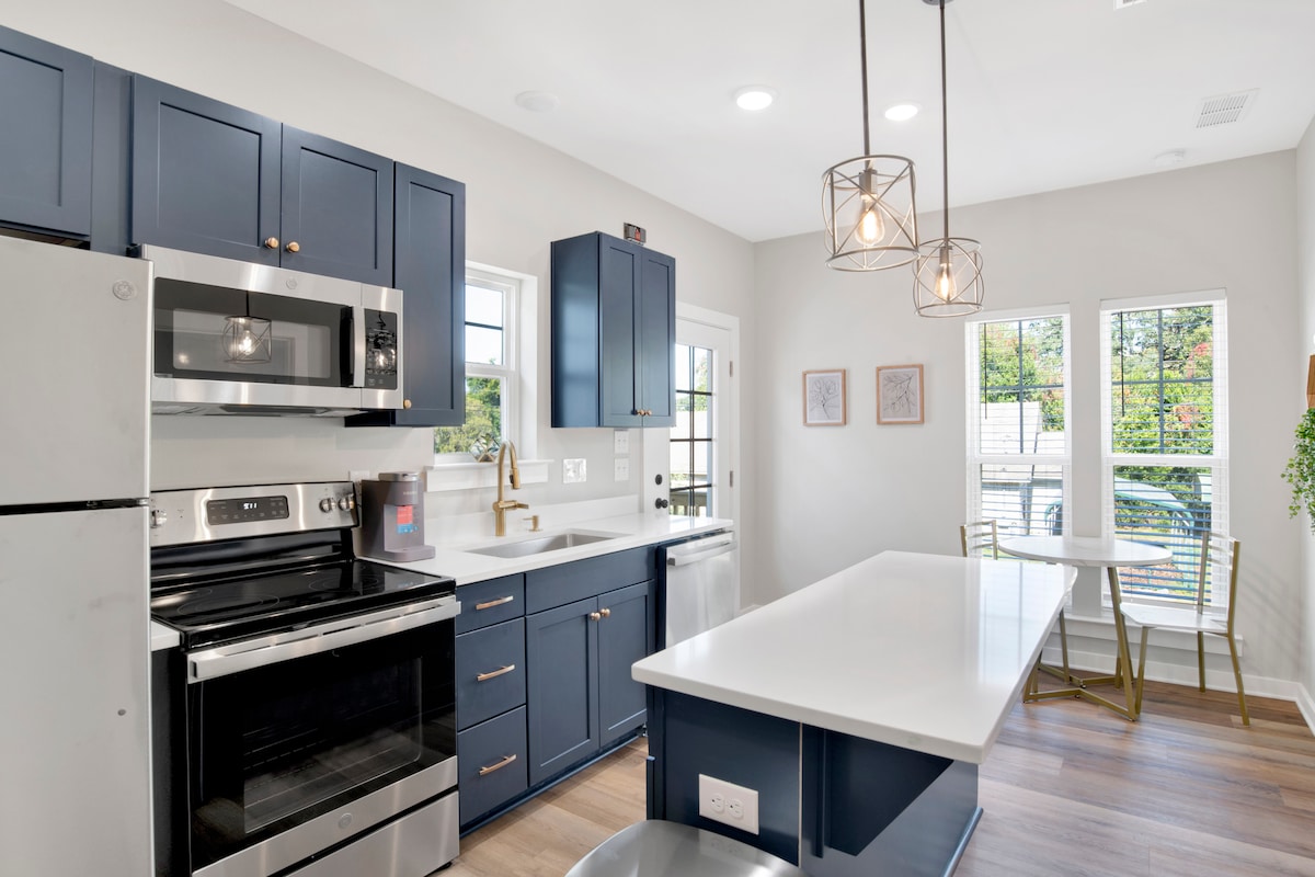 A contemporary kitchen features dark blue cabinetry paired with stainless steel appliances. A spacious island with a white countertop offers additional preparation space, while two windows allow natural light to illuminate the area. A small dining table with golden chairs is positioned nearby.