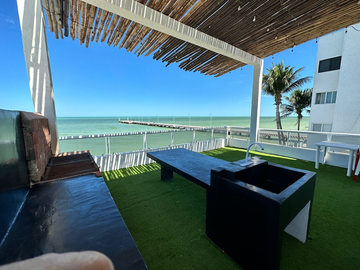 A spacious rooftop terrace offers a stunning view of the sea, with a black outdoor dining table and a built-in sink situated on green artificial turf. Overhead, a shaded bamboo canopy provides a relaxing atmosphere, while palm trees frame the picturesque coastal landscape.