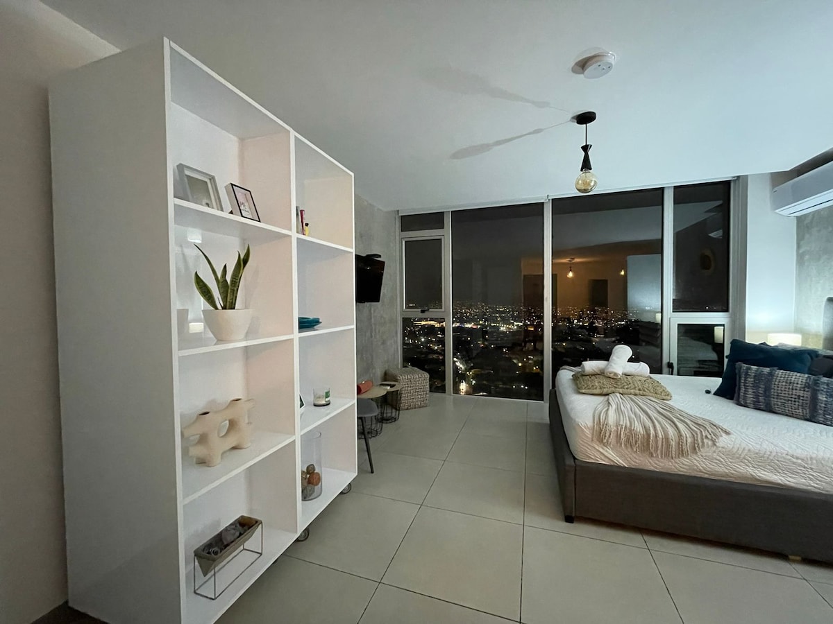 A modern living space is featured with a spacious bed and a large shelving unit displaying decor and plants. Floor-to-ceiling windows offer a stunning view of the city lights at night, enhancing the serene ambiance of the room.