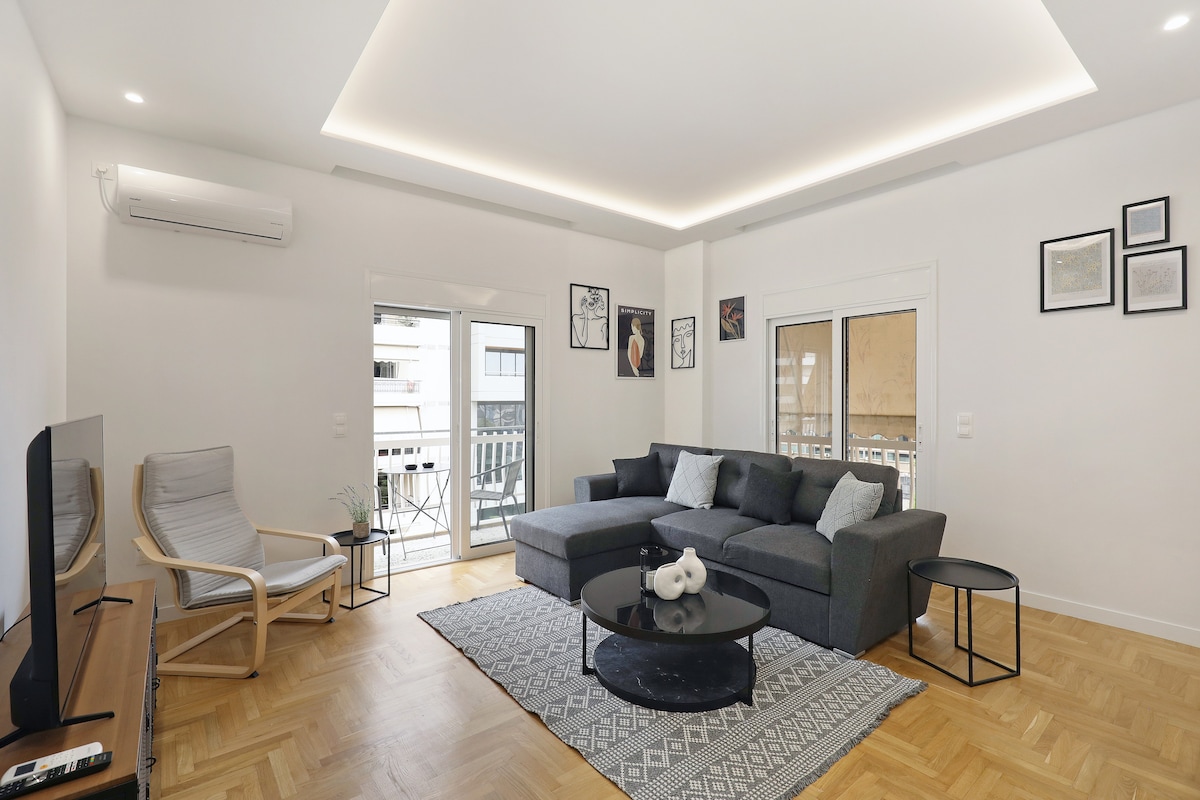 An open-concept living area features a spacious gray sofa and a small round table beside it. A light-colored armchair is positioned nearby, and a television is mounted on the wall. Natural light enters through two glass doors leading to a balcony, enhancing the airy feel.