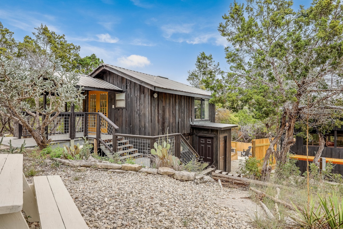 A multi-level cabin is showcased amidst natural landscaping, featuring a blend of wooden exterior and surrounding greenery. Stone walkways and a few native plants lead to the entrance, while the vibrant yellow door creates a welcoming focal point.