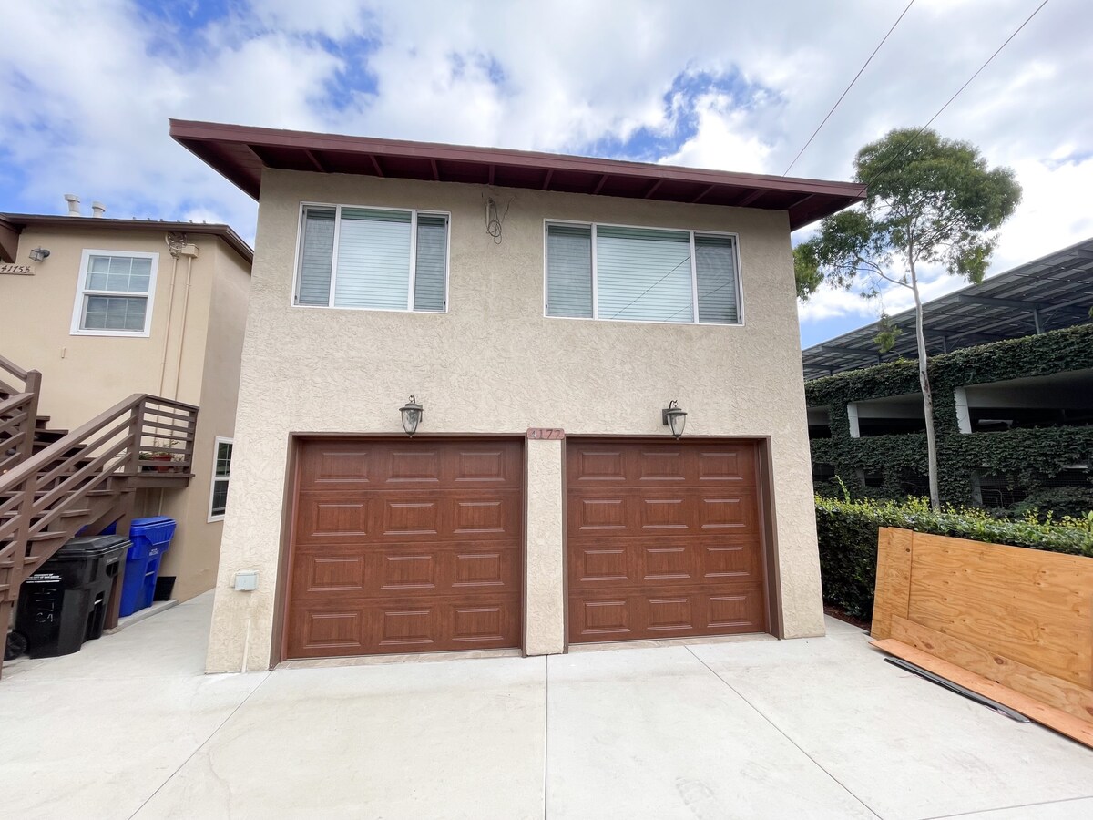 A two-story building is depicted, featuring a light-colored exterior with two spacious garage doors in a rich brown finish. Large windows are situated above the garages, allowing for natural light. Surrounding greenery and a neatly paved driveway are also visible.