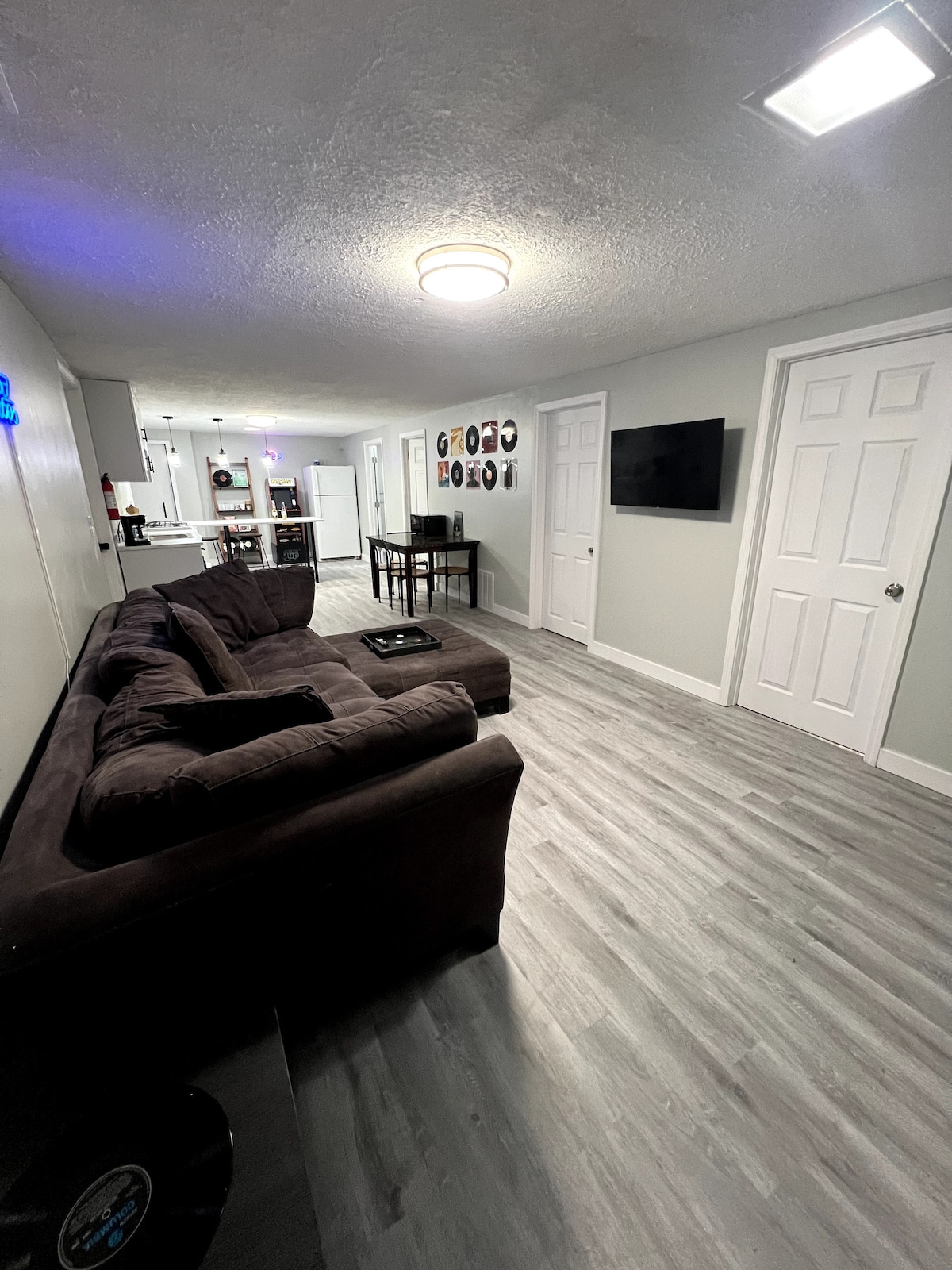 A spacious living area features a dark sectional couch facing a wall-mounted smart TV. Light-colored walls and flooring create a bright atmosphere. A small dining table is visible in the background, along with doors leading to additional rooms, offering a functional layout.
