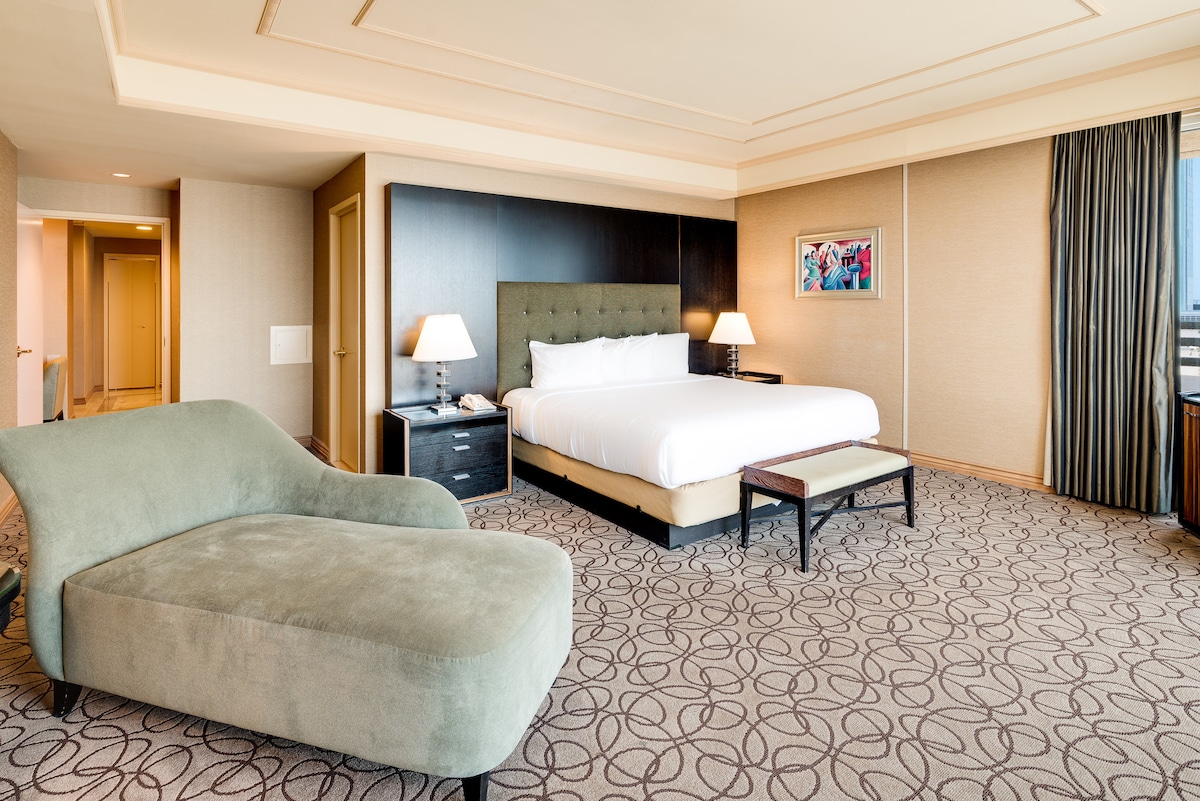 A spacious suite is showcased with a king bed and a large window allowing natural light. A comfortable sofa is positioned for relaxation beside a bedside table with a lamp. The neutral-toned decor and geometric patterned carpet create a serene environment.