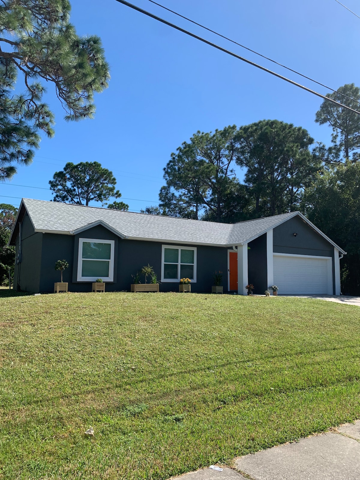 A single-story home is shown with a grey exterior and a welcoming orange front door. The property features a neatly maintained lawn and flower planters flanking the entrance. Tall trees in the background provide a sense of privacy and greenery.