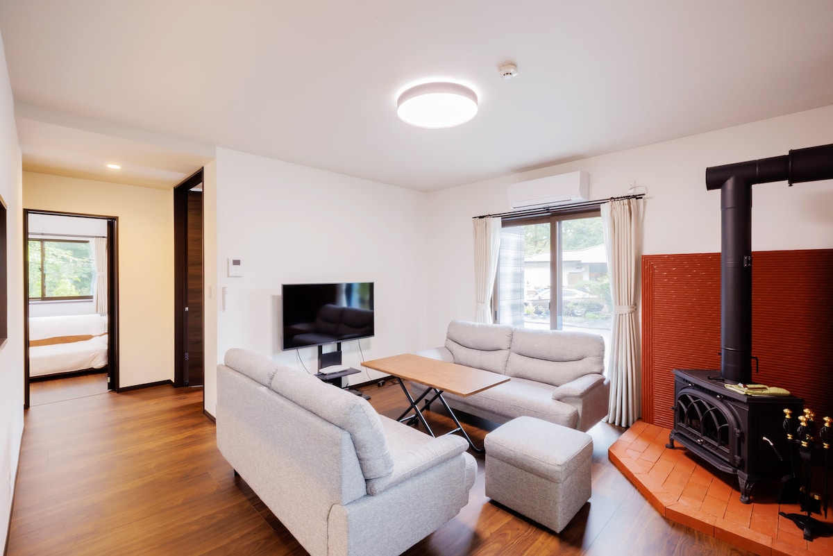 A spacious living area is furnished with two light gray sofas and a central table, promoting relaxation. A television is mounted on the wall opposite the seating. Natural light enters through a window beside a cozy wood stove, adding warmth to the inviting space.