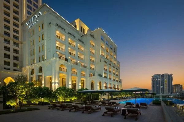 The exterior of Vida Residences is showcased at dusk, featuring modern architecture illuminated by warm lighting. Sunbeds line the pool area, nestled among lush greenery, creating an inviting space for relaxation. The pool reflects the vibrant city skyline and is framed by the elegantly designed building.