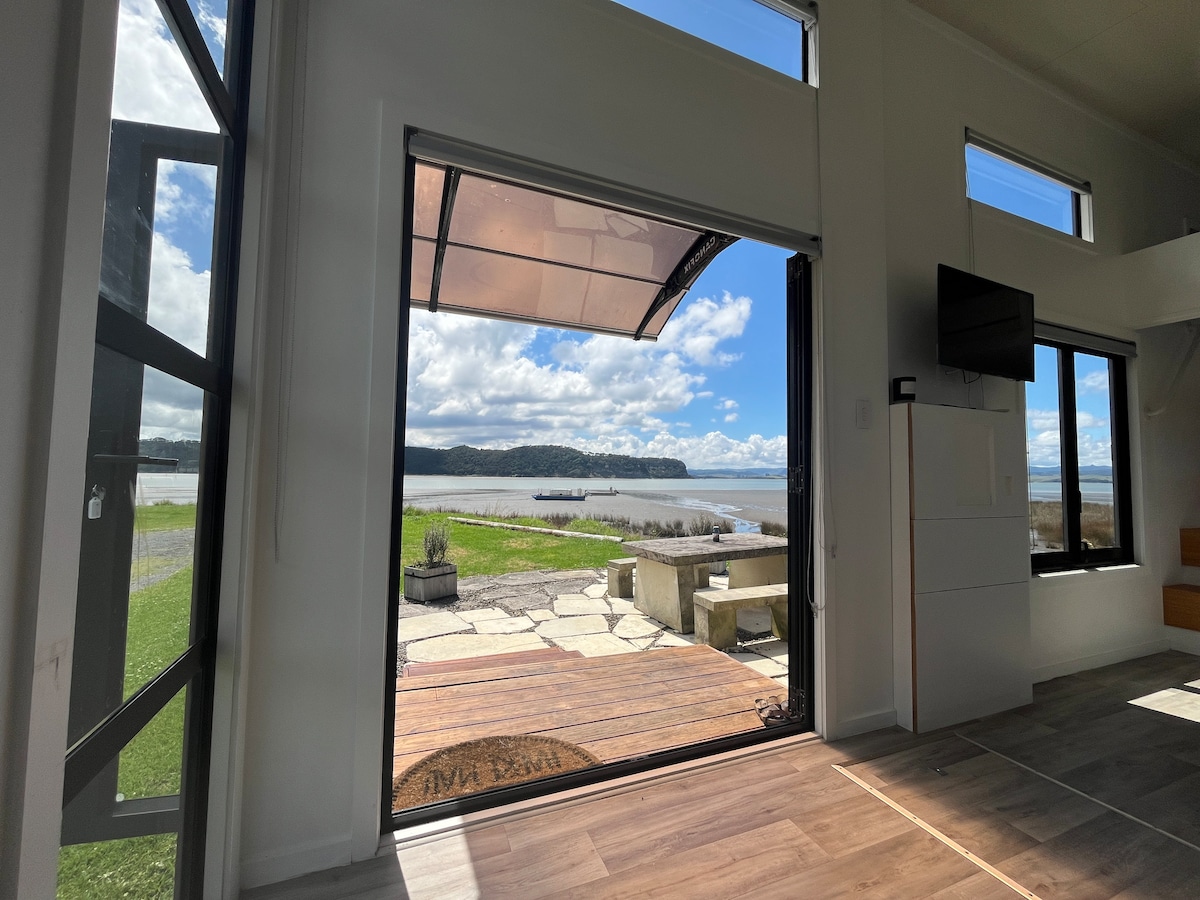 Expansive glass doors provide unobstructed views of Kawhia Harbour and the outdoor seating area. Natural light fills the living space, highlighting the stone patio and green lawn that lead down to the water's edge. Nearby islands and distant hills can be seen on the horizon.