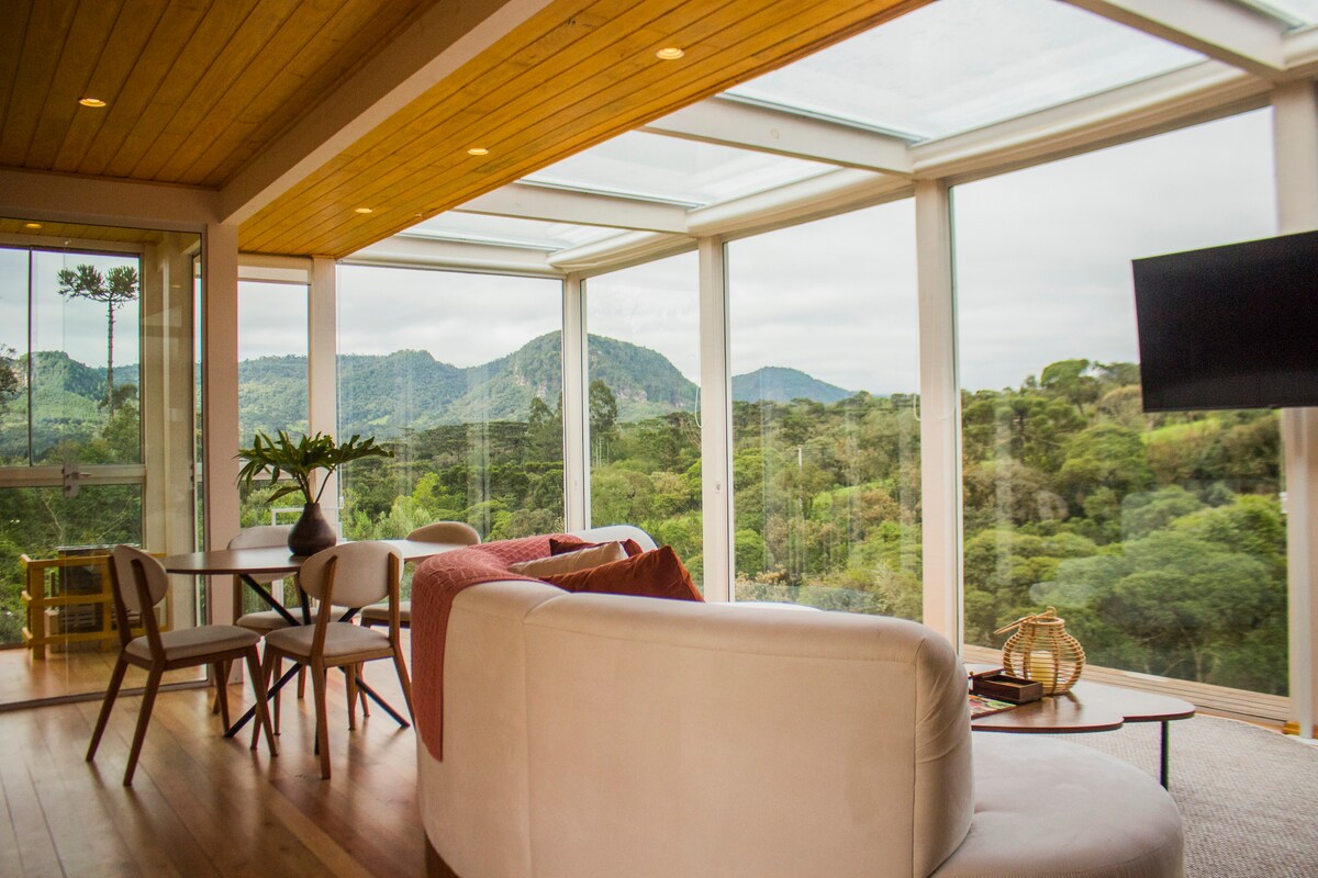 A spacious living area features a large glass wall that provides expansive views of the surrounding mountains and greenery. A comfortable white sofa is positioned with a small coffee table, while a dining table for four is set nearby, enhancing the connection to nature.