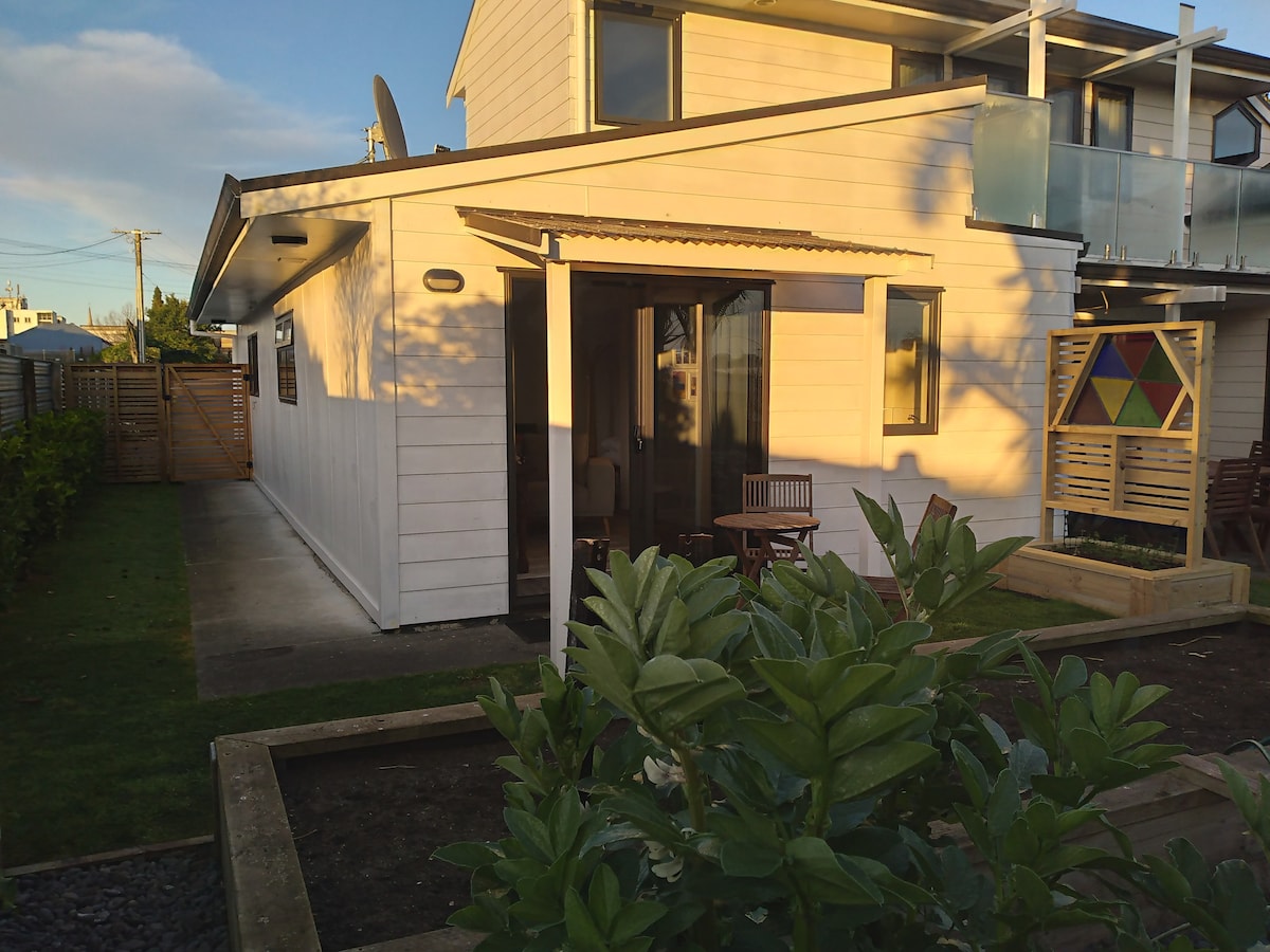 A side view of the studio displays a light-colored exterior with large glass doors leading to a small patio area. Garden beds with greenery frame the space, and a wooden table with chairs is positioned nearby, enhancing the outdoor living experience.