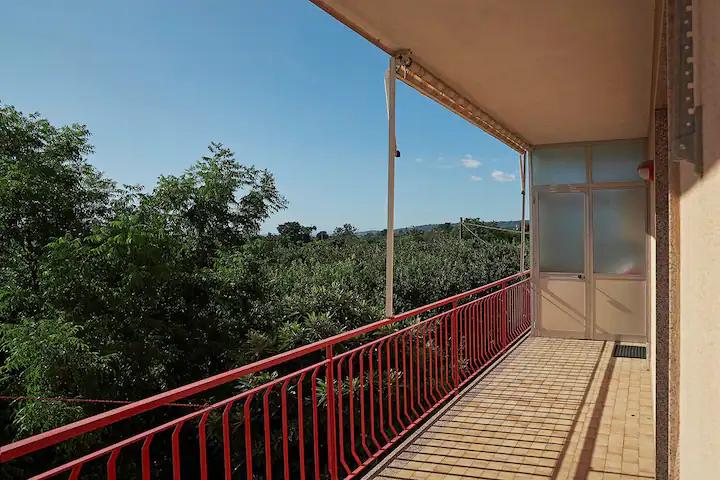 A private balcony is visible, featuring a red railing that offers a view of the surrounding greenery. Clear skies and distant trees create a serene backdrop. Sunlight casts patterns on the tiled floor, enhancing the outdoor space.