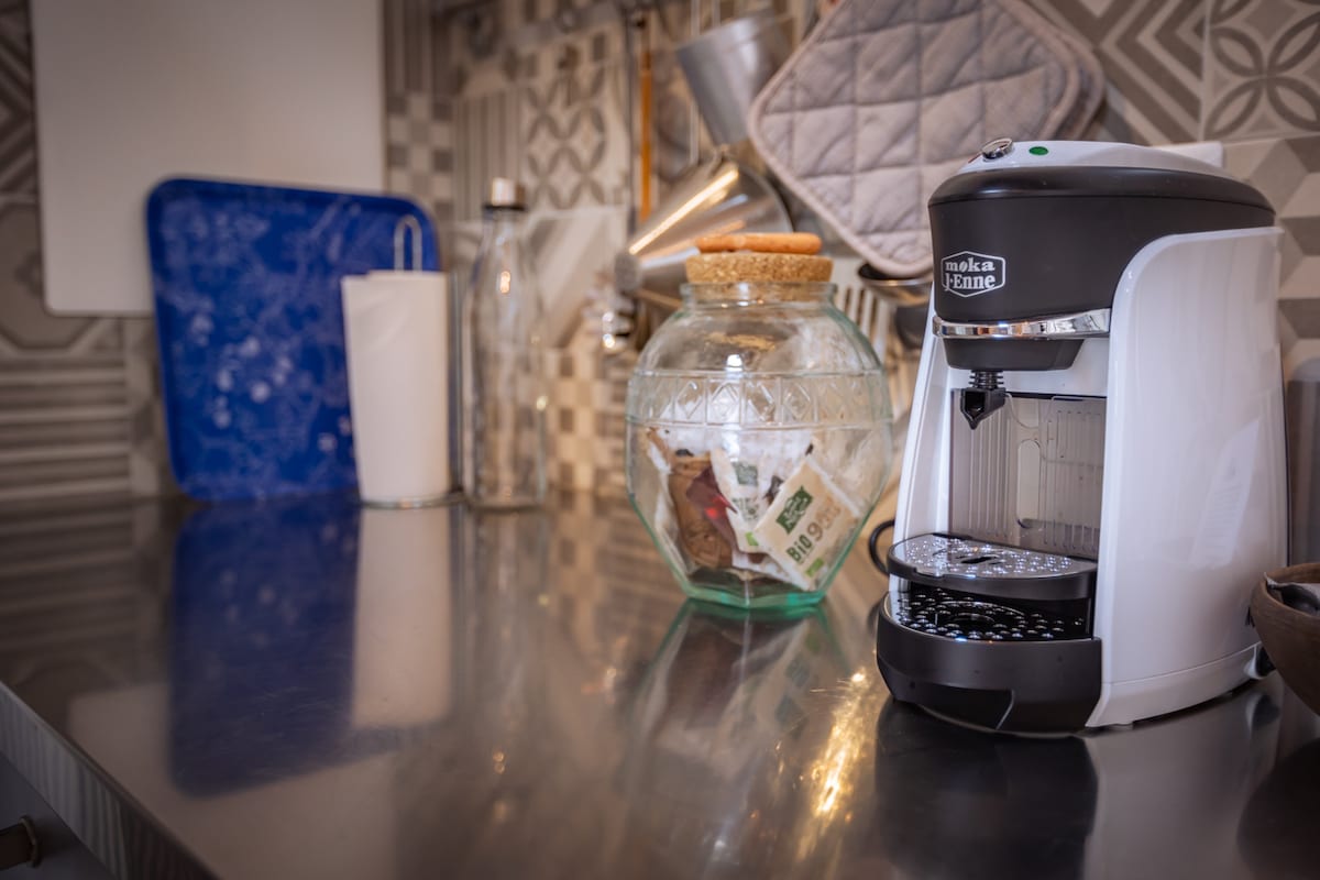A modern coffee maker is positioned on a sleek, stainless steel countertop. A decorative glass jar filled with various items is nearby, along with a white pitcher and a blue tray. The background features patterned tiles, enhancing the kitchen's contemporary design.