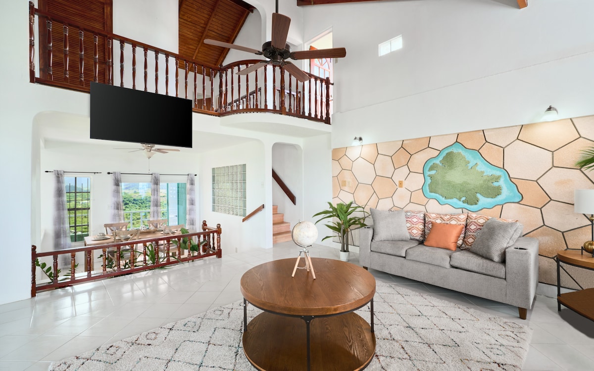 An airy living space features high ceilings and a large ceiling fan. A comfortable gray sofa is positioned near a round coffee table. A colorful wall art piece is displayed, and large windows offer views of the outdoors. A dining area is visible in the background.