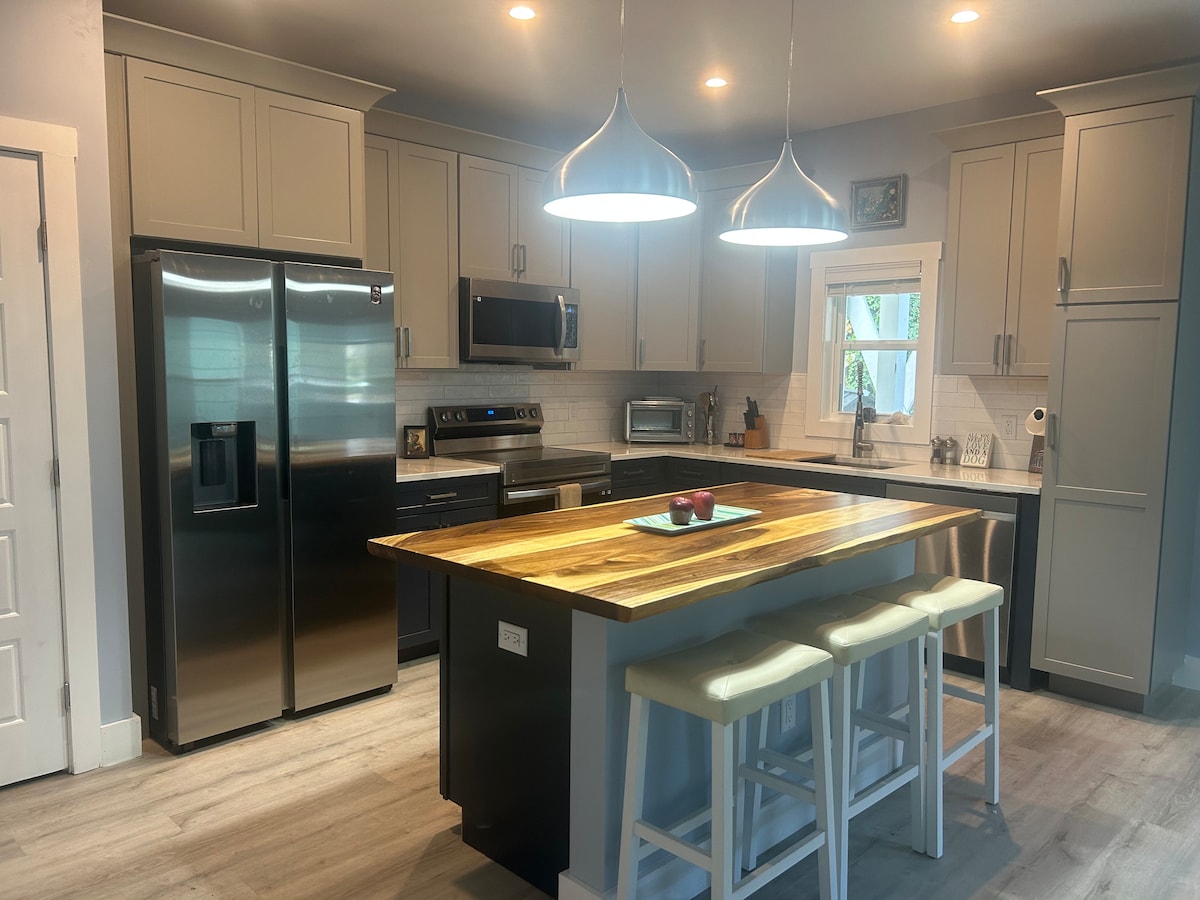 A modern kitchen features stainless steel appliances, including a refrigerator and microwave. A spacious island with a wooden countertop provides seating for three on light-colored stools. Under-cabinet lighting illuminates the sleek cabinetry and backsplash, creating a bright cooking space.