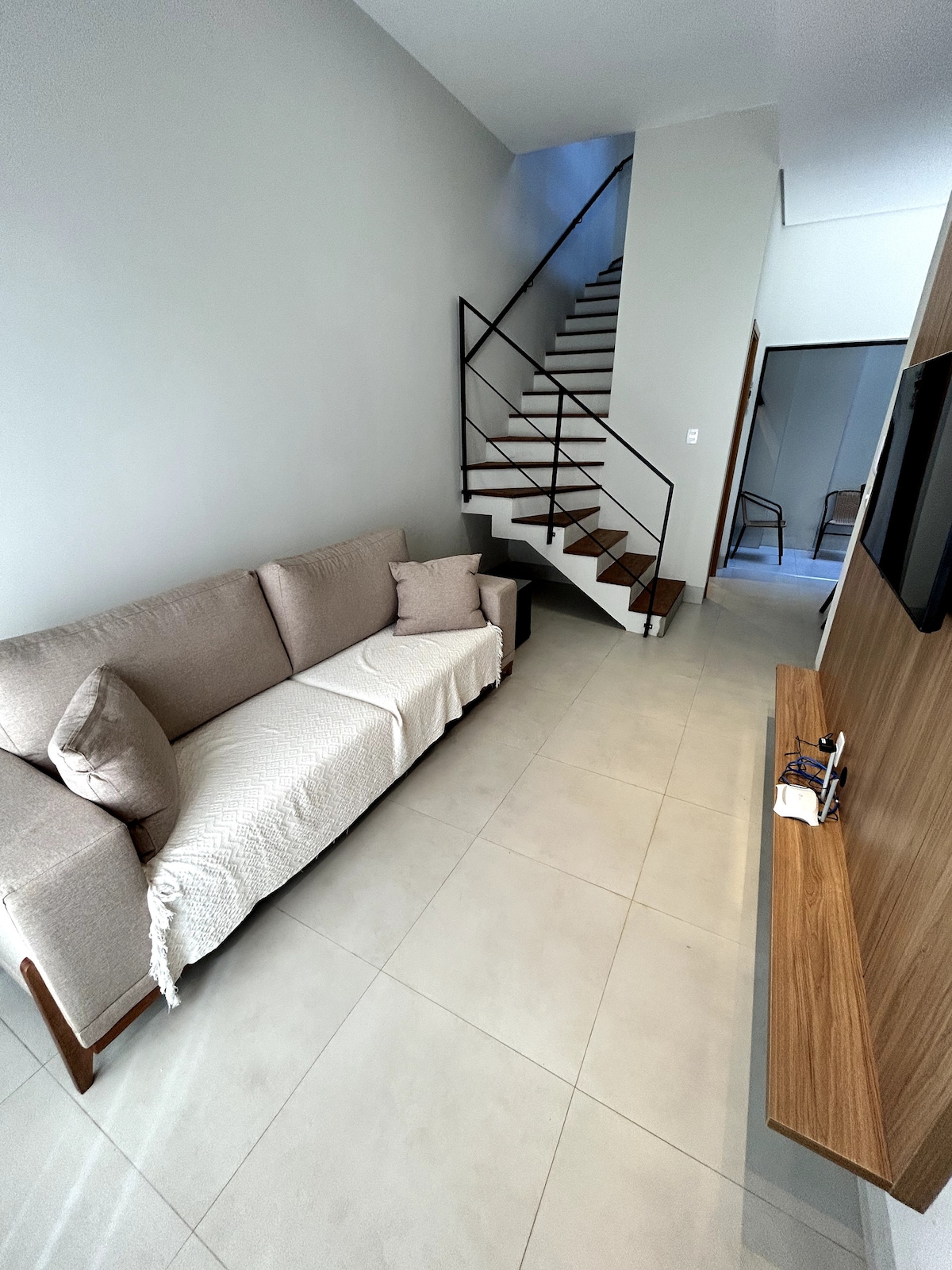 A light-filled living space is shown, featuring a neutral-toned sofa draped with a decorative throw. A modern staircase with metal railings leads to the upper level, complemented by sleek tile flooring that enhances spaciousness.