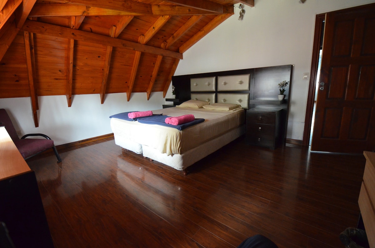 A cozy room features a spacious bed with neatly arranged linens and bath towels. The sloped wooden ceiling adds character, while wooden flooring enhances the warmth. A dark wooden nightstand is positioned beside the bed, and a chair is placed in one corner.