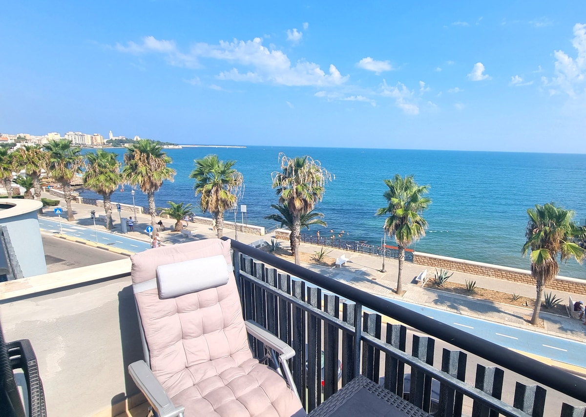 A spacious balcony overlooks the tranquil beachfront, featuring a comfortable lounge chair positioned for relaxing views of the sea and palm trees lining the shore. The clear sky enhances the serene atmosphere, inviting appreciation of the beautiful coastal landscape.