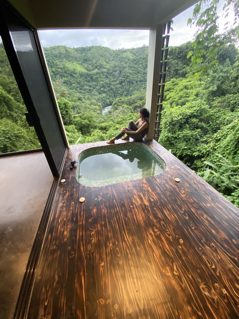 A private Jacuzzi is positioned on a wooden deck, overlooking dense tropical greenery and hills. Large glass doors provide a seamless transition between indoor and outdoor spaces. A person sits nearby, enjoying the serene view of the lush landscape and distant river.