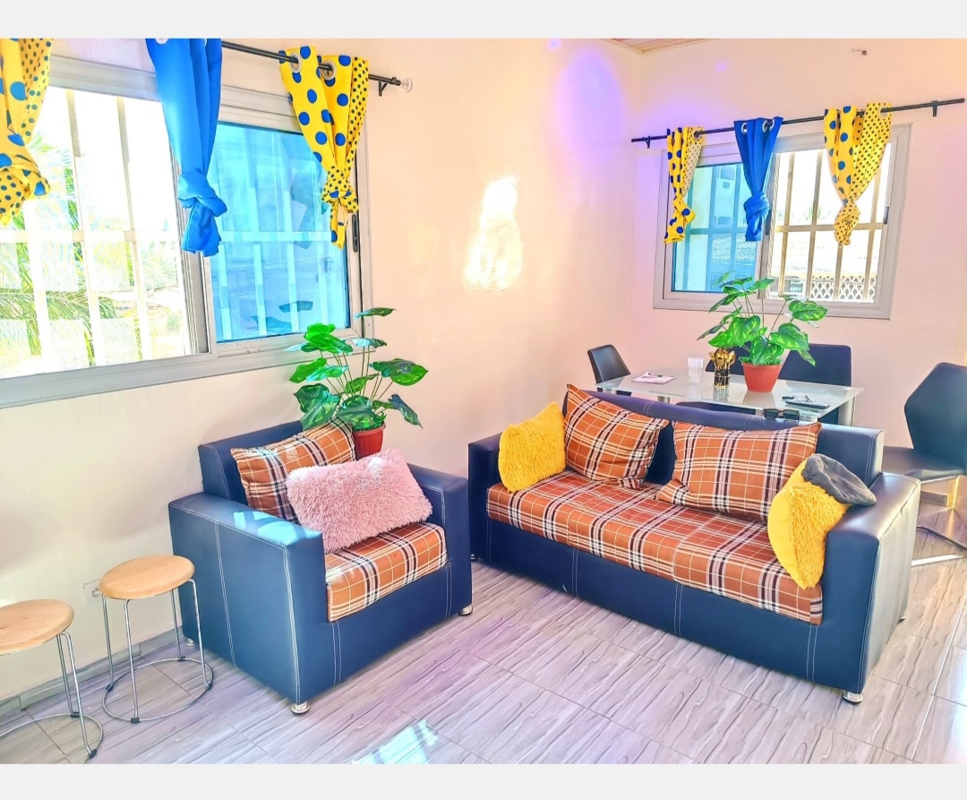 A stylish seating area features a comfortable two-piece sofa set in blue with plaid upholstery. Bright yellow cushions add color, while potted plants enhance the space. Natural light filters through large windows, complemented by vibrant curtains, creating a cheerful ambiance.