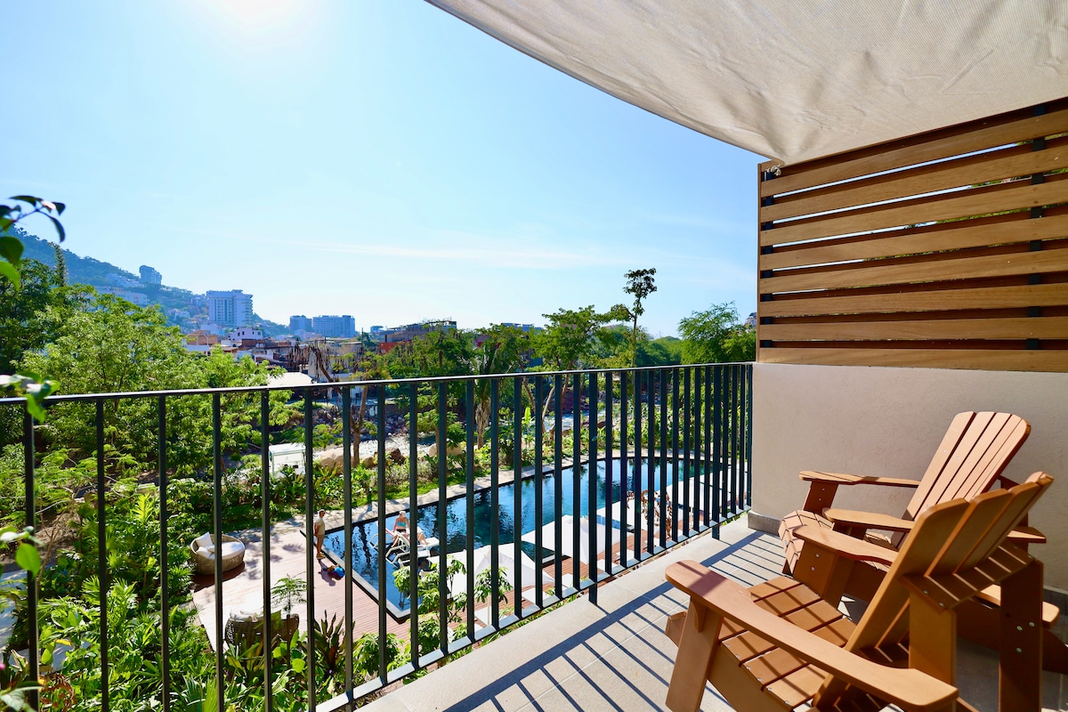 A private balcony is featured with wooden Adirondack chairs overlooking lush greenery and a pool area below. The scene is bathed in natural light, framed by a partially shaded awning, highlighting the serene outdoor space.