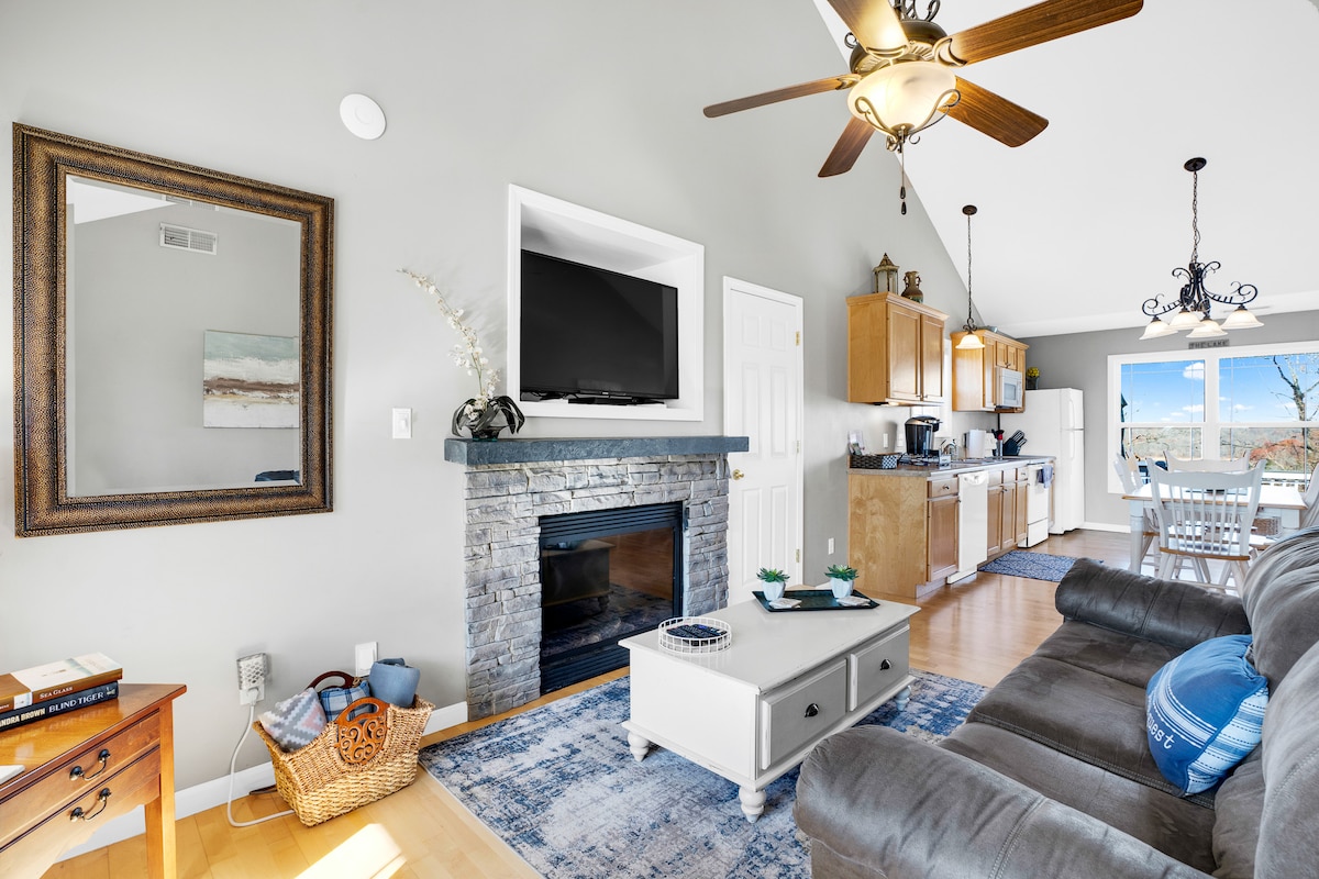 A welcoming living area is featured, showcasing a comfortable sofa and a stone fireplace beneath a flat-screen television. Light wood cabinetry lines the kitchen area, while dining space is visible through large windows that frame views of the surrounding landscape.