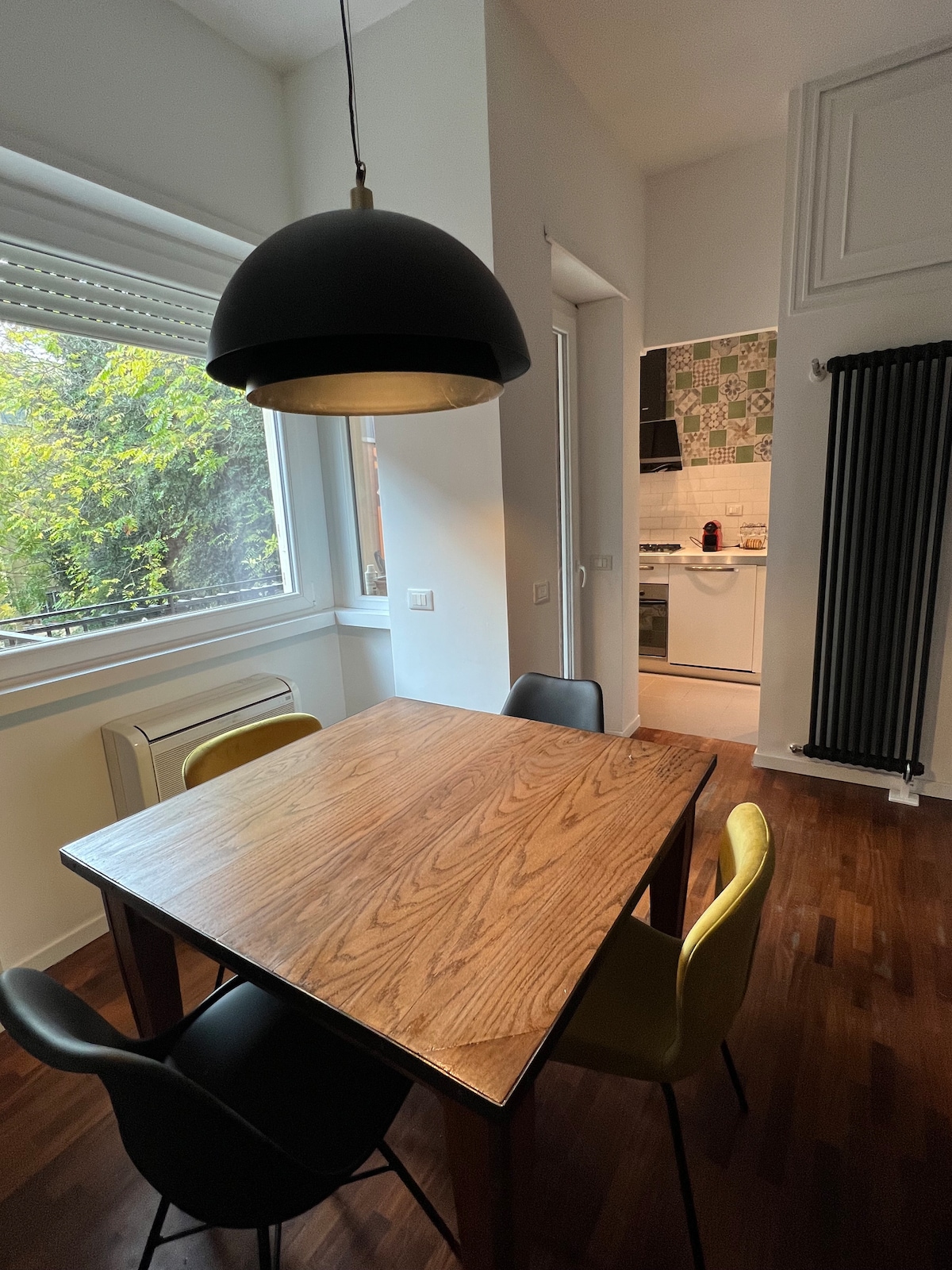 A dining area features a wooden table surrounded by four chairs in varying colors. Natural light streams through a large window with greenery visible outside. A modern pendant light hangs overhead, while a glimpse of the kitchen can be seen in the background.