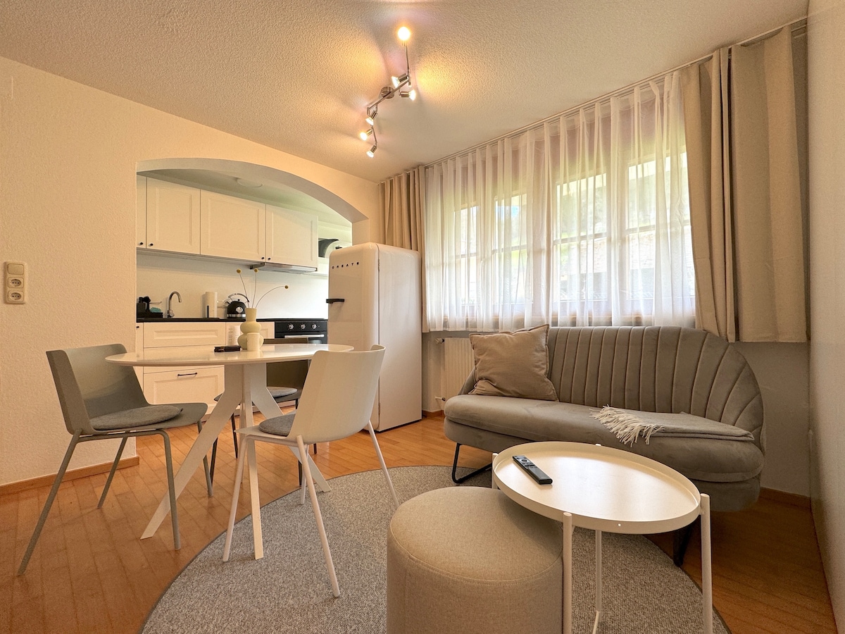 A cozy living area features a modern curved sofa and a circular coffee table. Light wooden flooring complements the pale walls, while large windows allow ample natural light to illuminate the space. A dining table with chairs is positioned nearby, creating an inviting atmosphere.