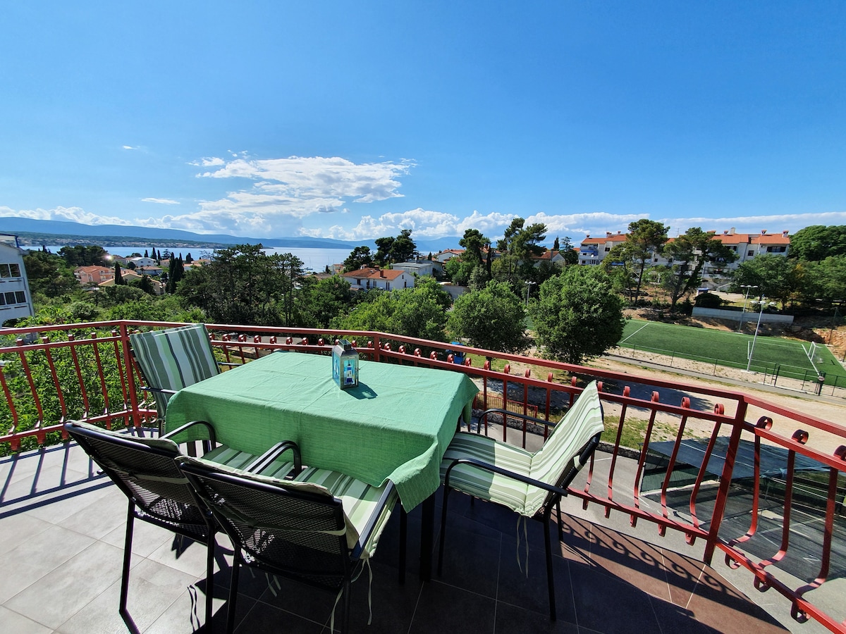 A balcony features a green tablecloth covering a table, surrounded by two chairs with striped cushions. The view overlooks a landscaped area with trees and greenery, and a distant view of the sea is visible under a bright blue sky.