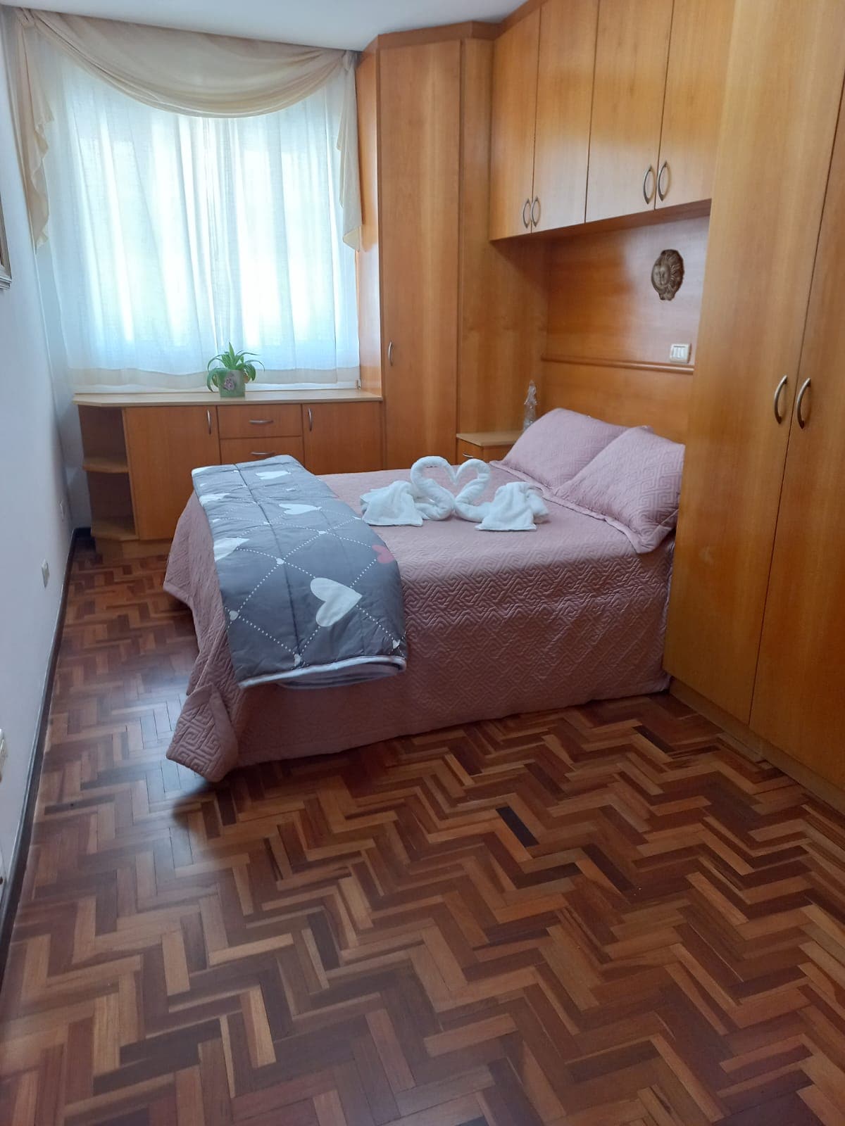 A well-appointed sleeping area features wooden cabinetry and a spacious bed adorned with soft linens. Light filters through a large window, enhancing the warm wood tones of the herringbone floor. A small plant decorates the bedside table, adding a touch of greenery to the room.