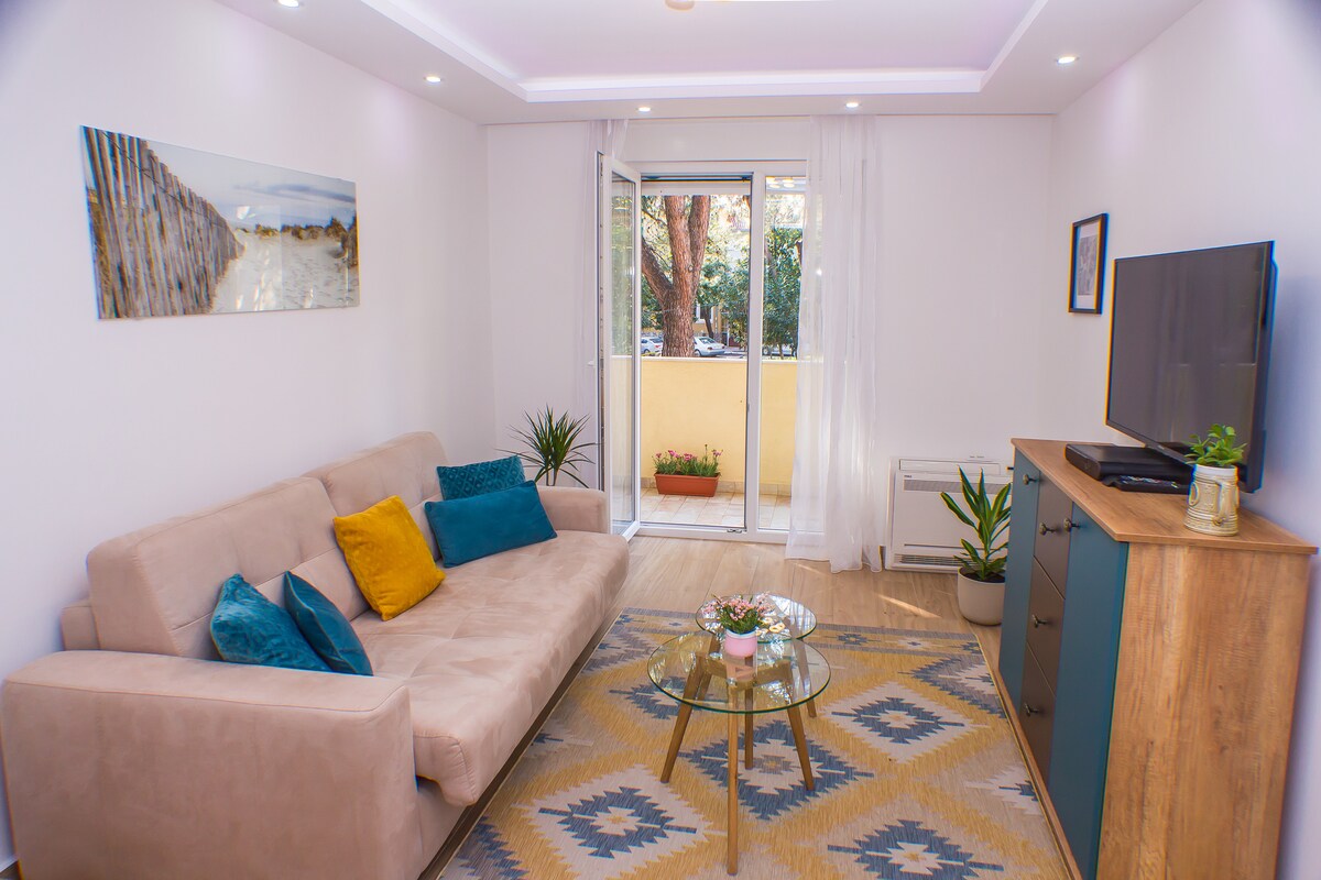 A comfortable seating area features a light-colored couch adorned with colorful cushions. A glass coffee table sits on a patterned rug beside a flat-screen TV. Natural light filters through the sliding doors, which open to a small balcony with potted plants.