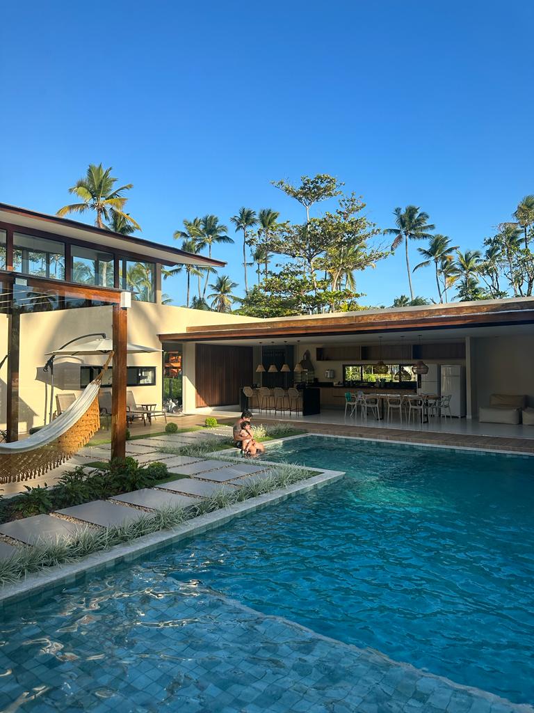 An inviting outdoor space showcases a tranquil pool bordered by tropical greenery. A shaded gourmet area is visible behind the pool, with seating and a view of the nearby palm trees. A hammock adds a relaxing touch to the serene environment.