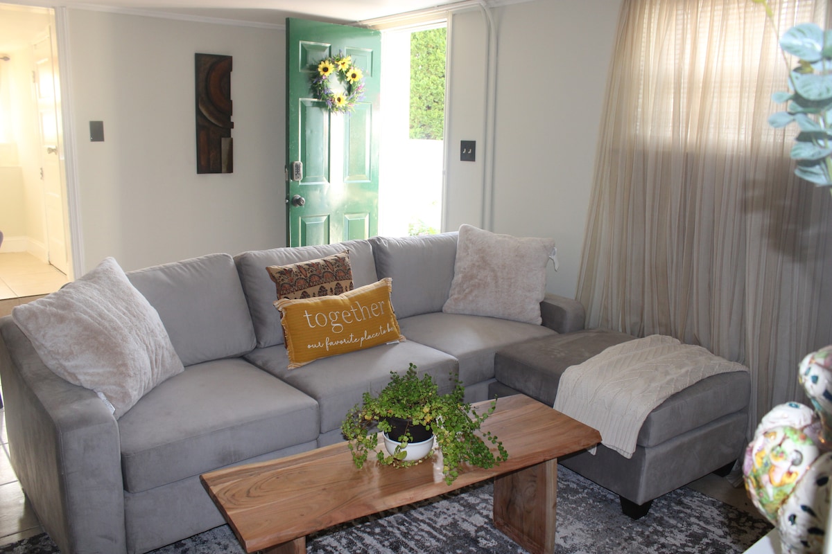 The living area features a gray sectional sofa adorned with plush throw pillows. A wooden coffee table with a potted plant adds a natural touch. Light drapes softly through sheer curtains, and a vibrant green door complements the welcoming atmosphere.