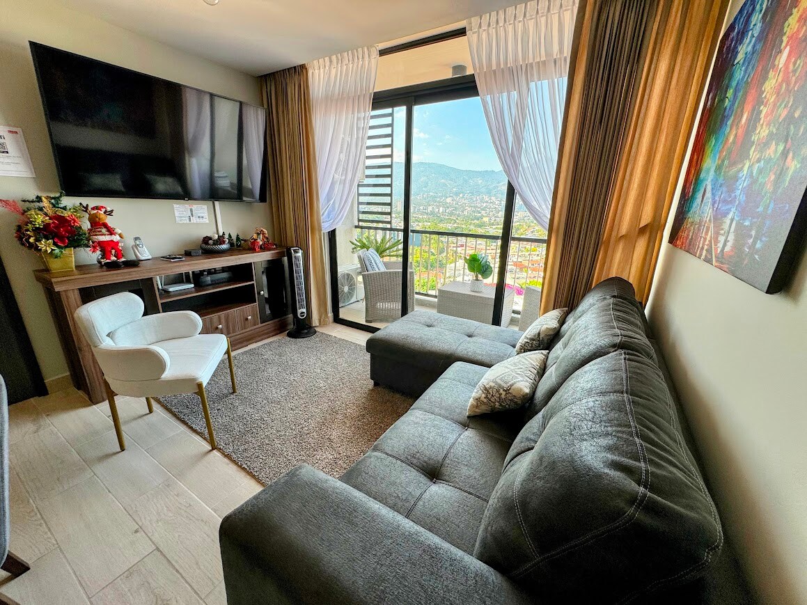 A modern living area features a plush sectional sofa and an armchair on a soft area rug. A large TV is mounted on the wall, and expansive glass doors reveal a stunning city and mountain view, framed by light curtains.