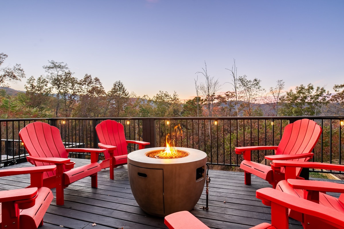 A circular firepit is surrounded by six red Adirondack chairs on a wooden deck. The scene captures the evening twilight, with distant trees in fall colors, creating a serene atmosphere for gatherings.