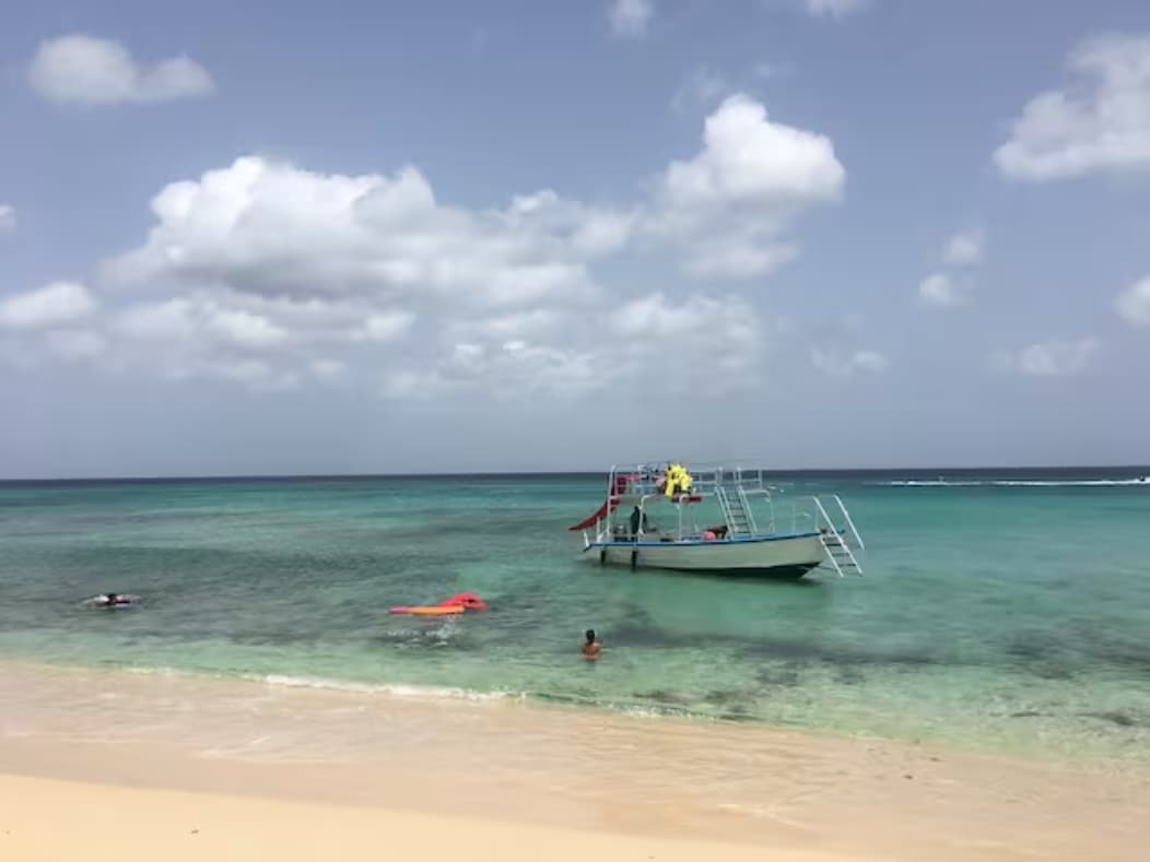 A sandy beach extends along the shoreline, where a boat is anchored in clear turquoise waters. Swimmers are seen enjoying the calm sea, with gentle waves lapping against the shore under a partly cloudy sky.