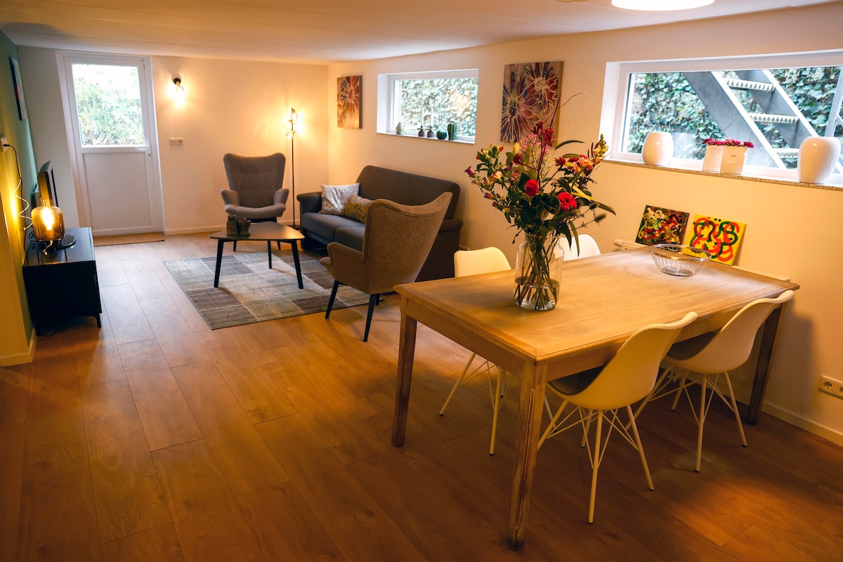 A spacious living area is highlighted by large windows that allow natural light to fill the room. A comfortable gray sofa and a pair of armchairs invite relaxation. A wooden dining table with white chairs adds to the welcoming atmosphere, complemented by decorative art pieces on the walls.