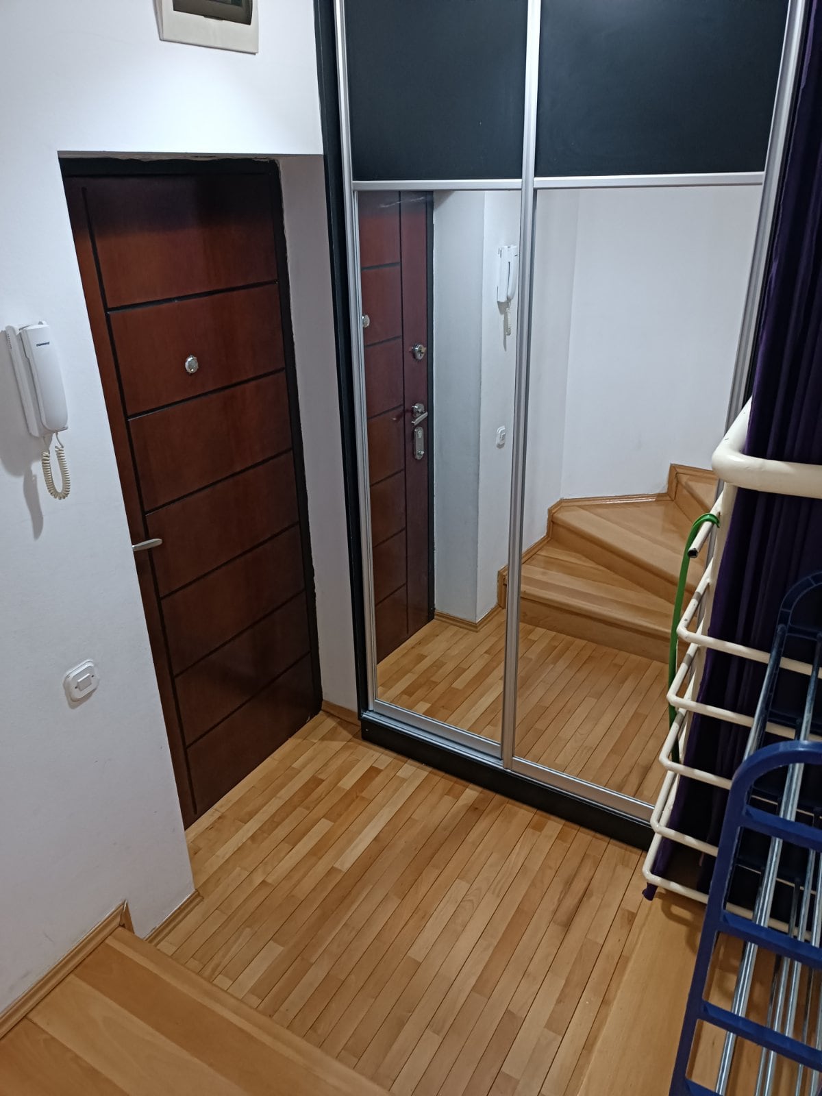 A well-lit entryway features wooden flooring and a set of stairs leading upward. A full-length mirror reflects the space. The entrance door and a mirrored closet door are visible, along with a wall-mounted intercom handset.