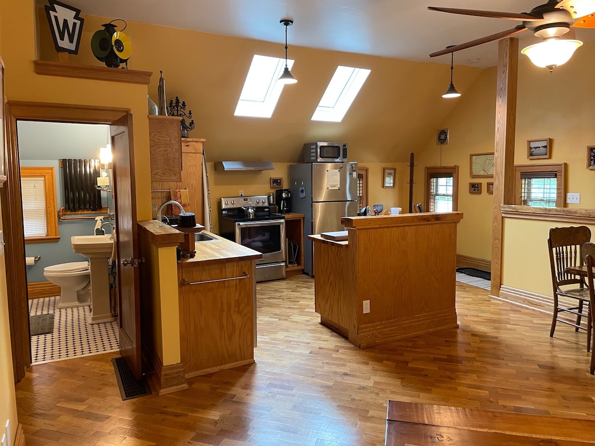 The open kitchen and living area features high ceilings and natural light from skylights. Wooden cabinetry and a spacious island create a functional cooking space. A dining table is present, and in the background, a bathroom door is visible, leading to additional amenities.