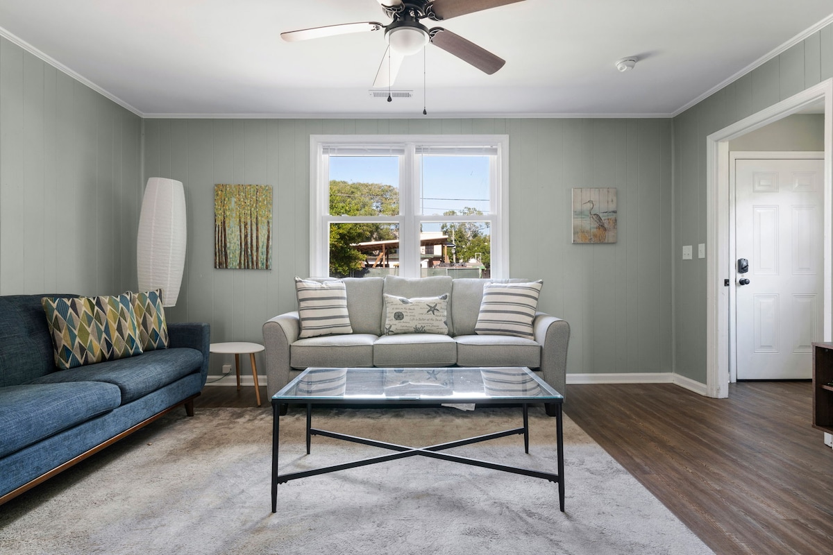 The living area features a comfortable grey sofa paired with a blue accent chair. A glass coffee table is centered on a light-colored rug, and two windows allow natural light to fill the space. Wall art adds subtle color to the neutral walls.